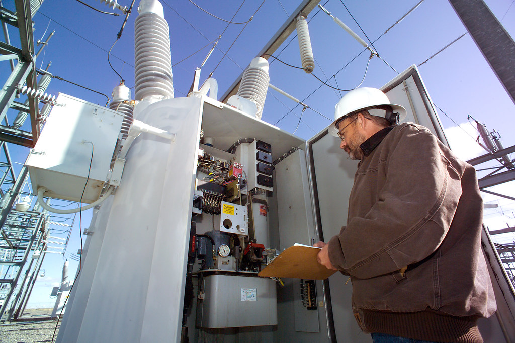 A technician in a hard hat inspecting electrical switchgear at a substation